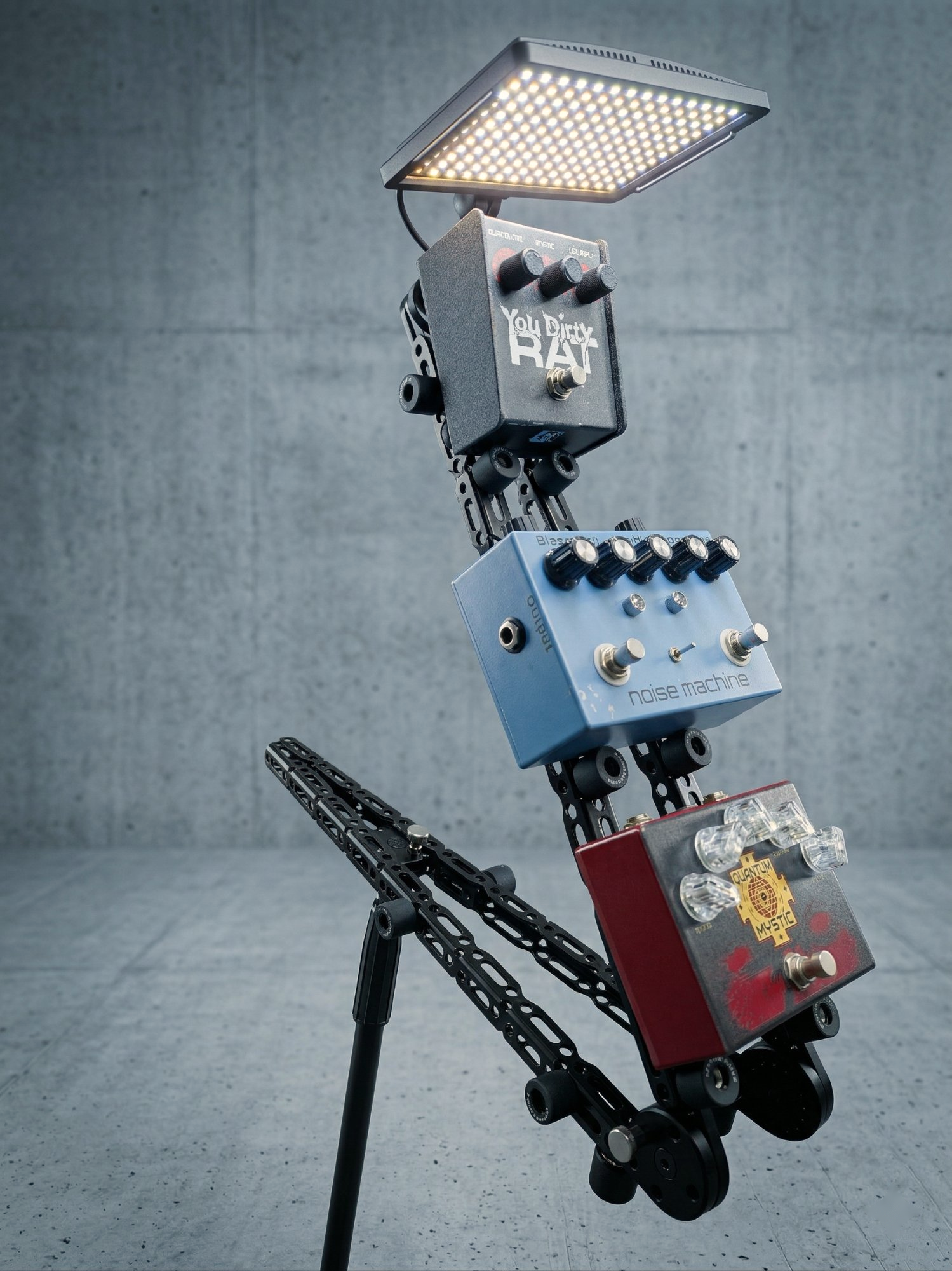 Three stompbox effects pedals, You Dirty RAT distortion, Blackout Effectors Noise Machine, and Quantum Mystic, mounted on a Manofold at standing height on a mic stand alongside an LED panel, in a concrete studio setting.