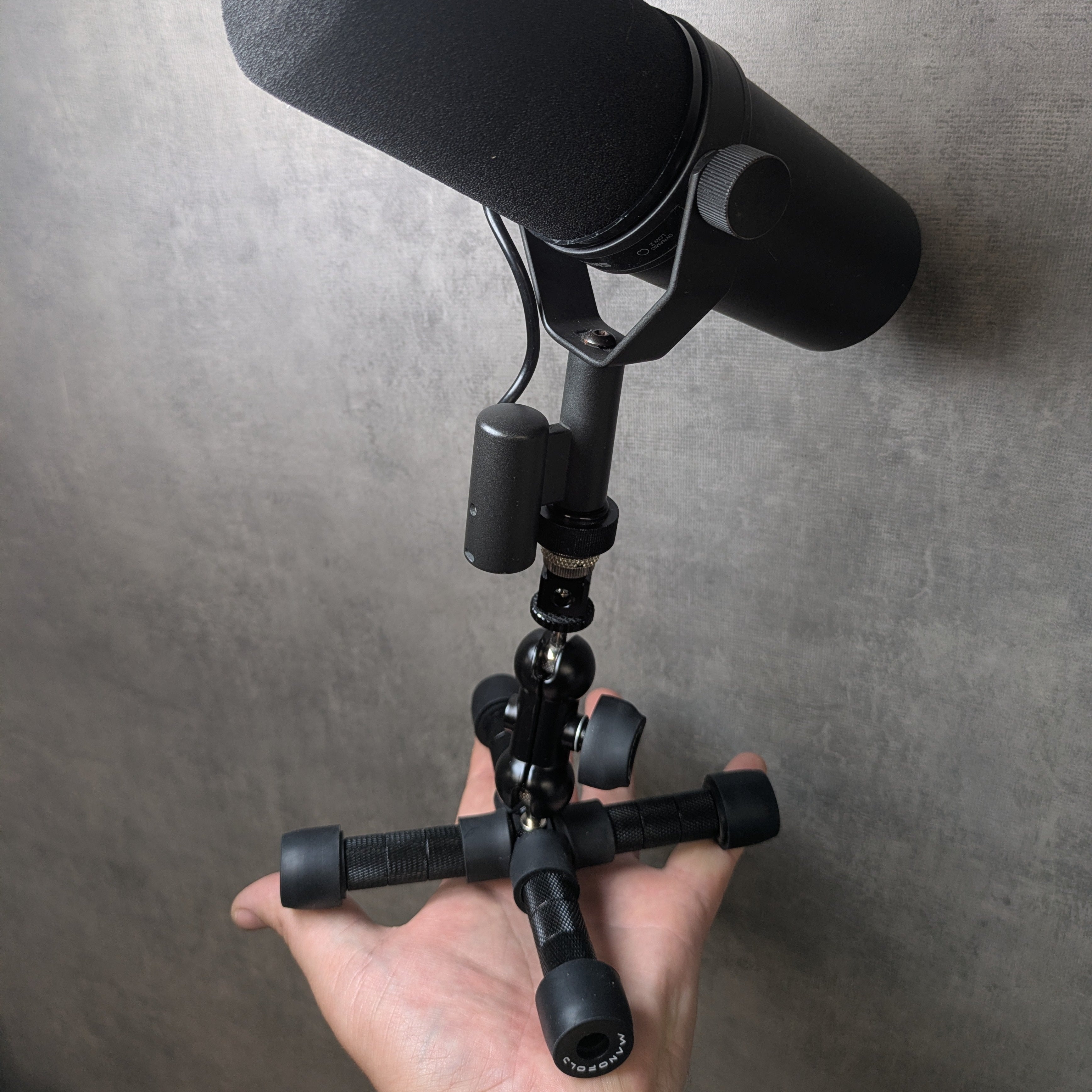 A black studio microphone with a foam windscreen, mounted on a small black Manofold stand, held in a person's hand against a concrete.